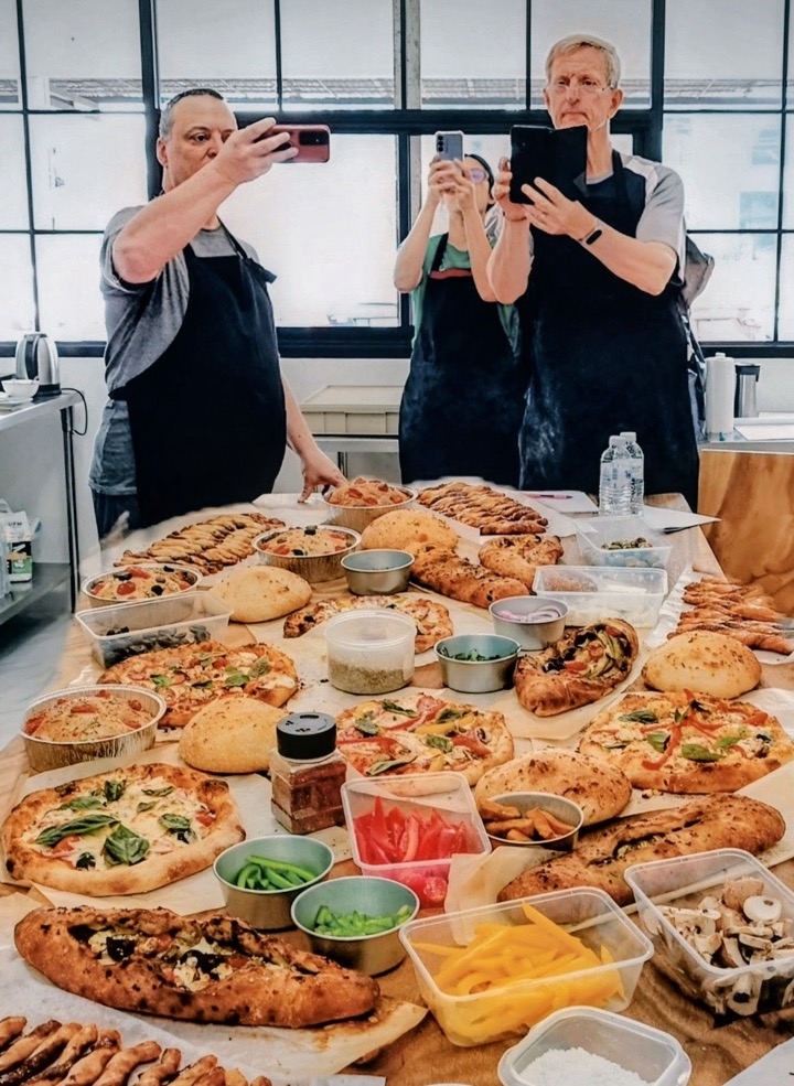 Class guests in aprons taking photos of a long table full of the pizzas, focaccia and breads they baked