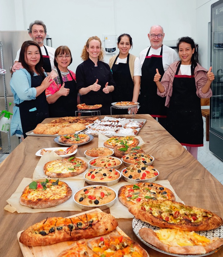 A class group giving thumbs up behind a table full of homemade pizzas and flatbreads