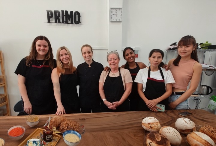 A class group in aprons smiling under the PRIMO sign, with their sourdough loaves on the table